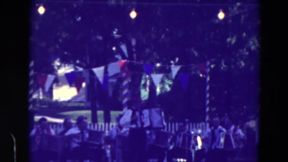 1965 Clarkson, Nebraska home-movie frame showing accordion players and spectators near a white fence beneath patriotic bunting and bulbs.