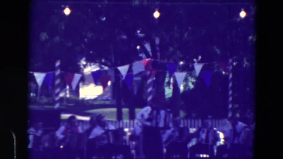 Blue-tinted 8mm frame of a Clarkson, Nebraska festival accordion group performing outdoors with bunting overhead and people seated nearby.