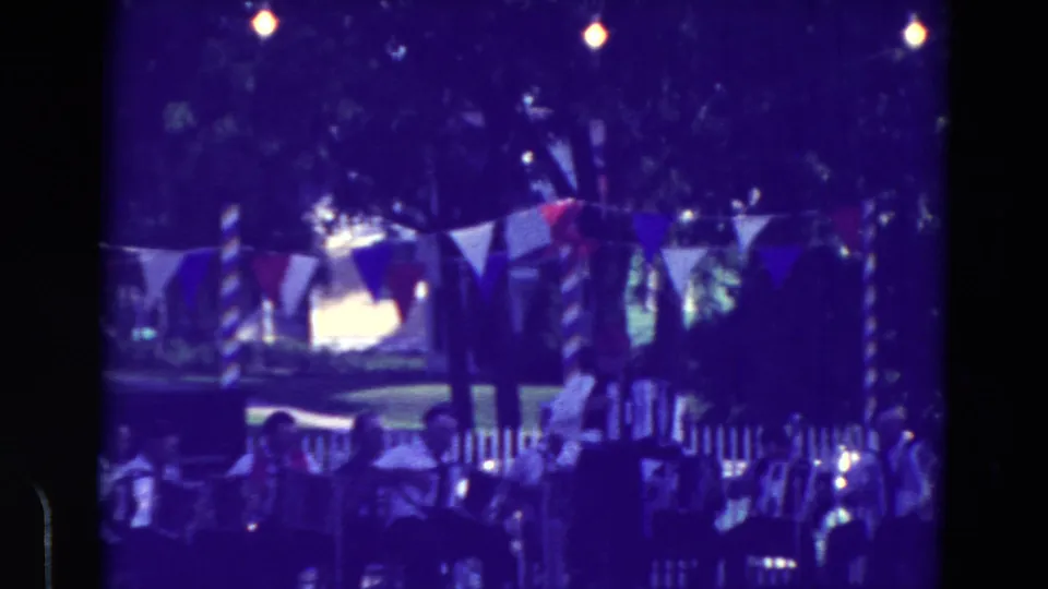 Home-movie still of the community festival performance, with accordion players partly silhouetted under bunting and string lights in Clarkson, Nebraska.