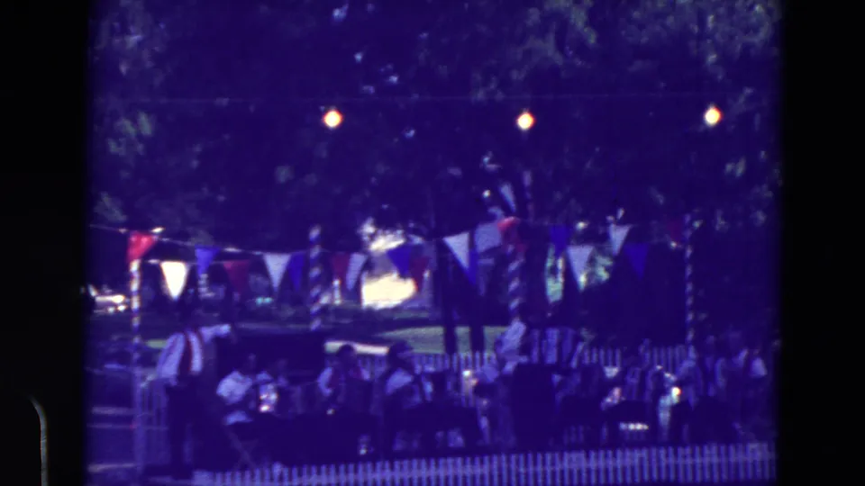 Wider 1965 Clarkson, Nebraska festival frame showing accordion musicians and seated viewers behind a white fence under red, white, and blue bunting.