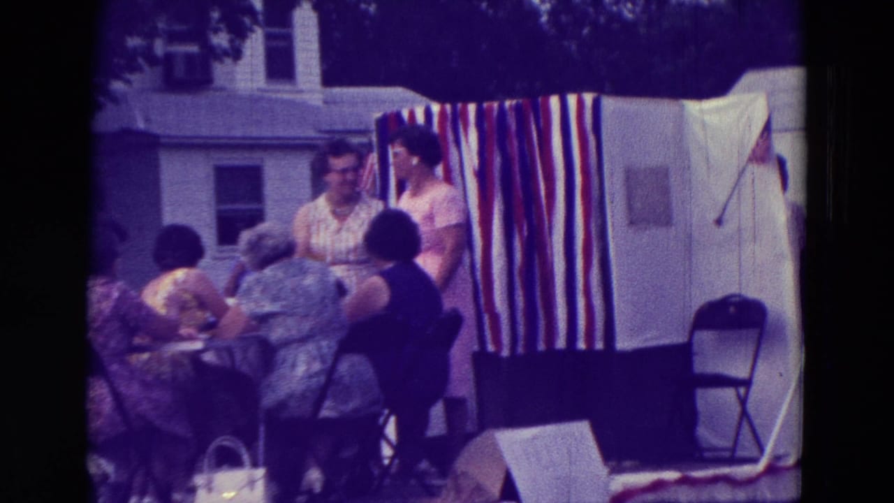 Women gather beside a patriotic striped parade float near a small-town street.