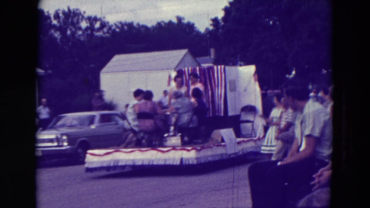 A decorated float carrying people passes spectators and parked cars on a residential street.