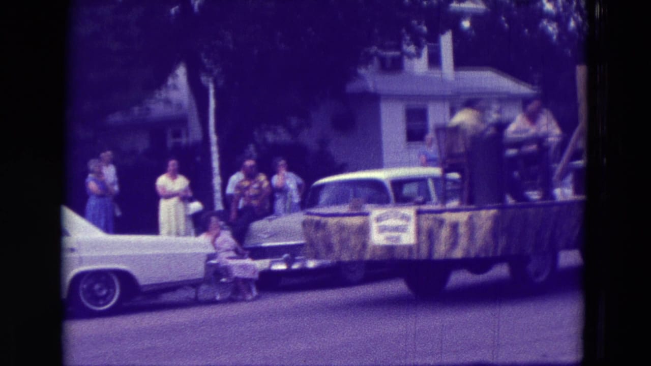 1965 Clarkson, Nebraska parade view with a light blue sedan, decorated float, spectators, houses, and trees along the street.