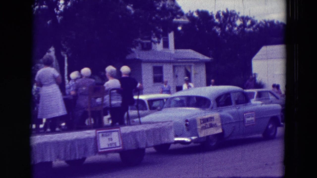 People stand on a parade float while a classic car is parked or moving beside it.