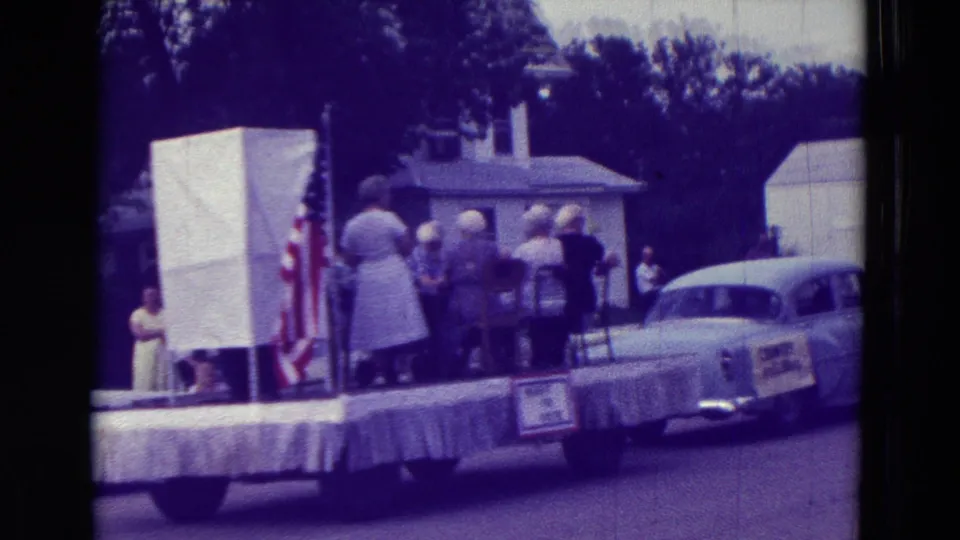 Parade float in Clarkson, Nebraska, with adults seated and standing near signs, an American flag, and a classic car nearby.