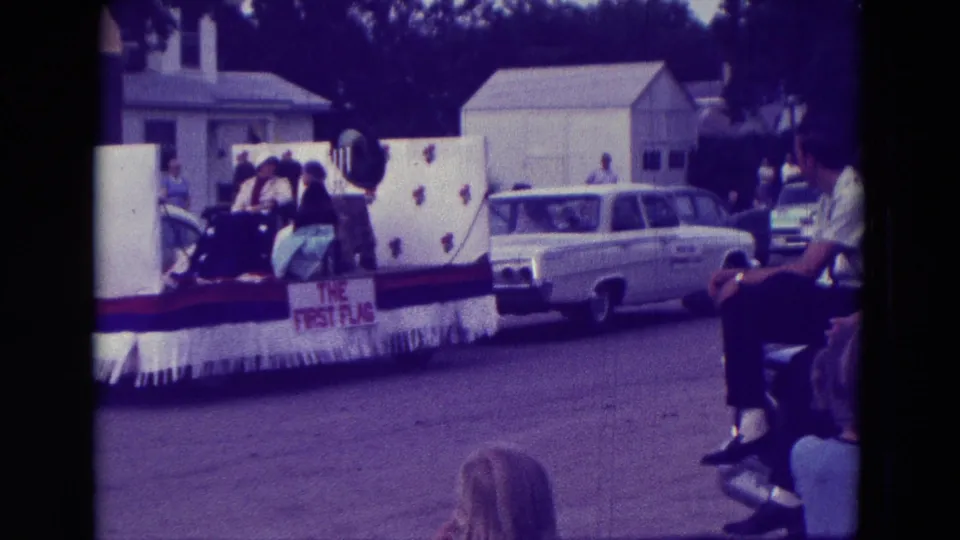 A decorated float with white panels and visible text passes spectators and a station wagon during the 1965 Clarkson parade.