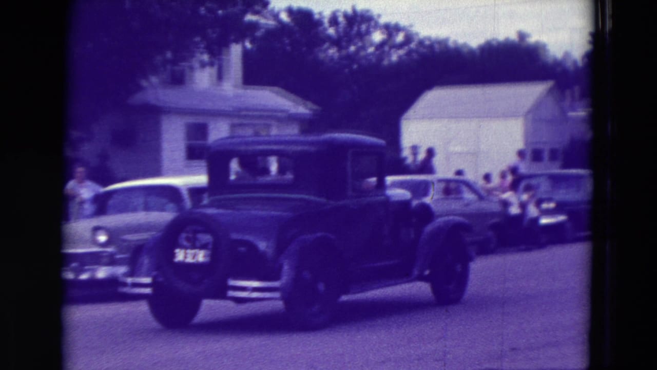 Black sedan driving past a white car, houses, and spectators.