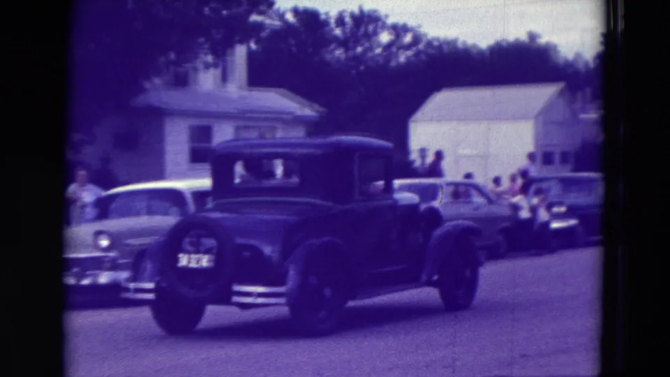 A black vintage sedan passes a white car and roadside spectators in the 1965 Clarkson, Nebraska street footage.