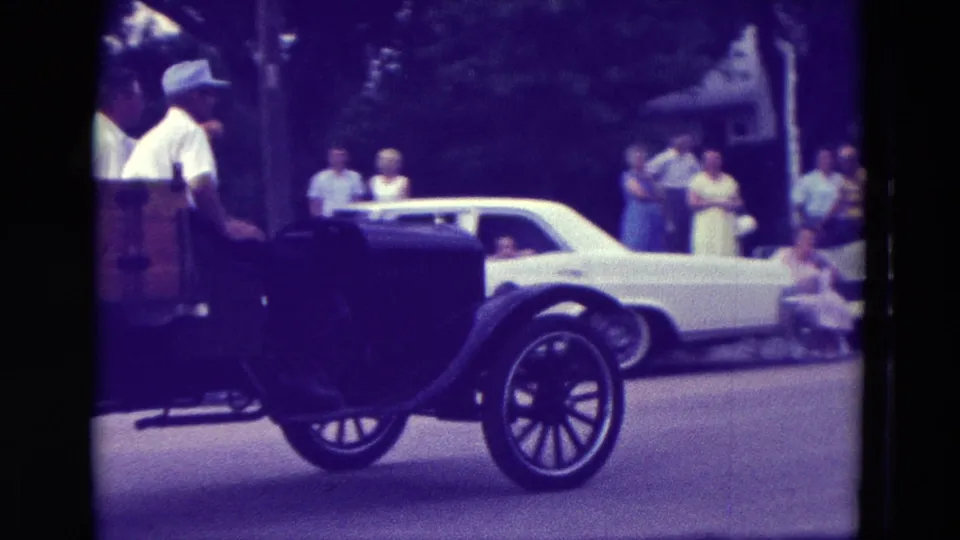 A dark open vintage vehicle passes in front of spectators and a white car in the Clarkson, Nebraska 1965 home-movie scene.