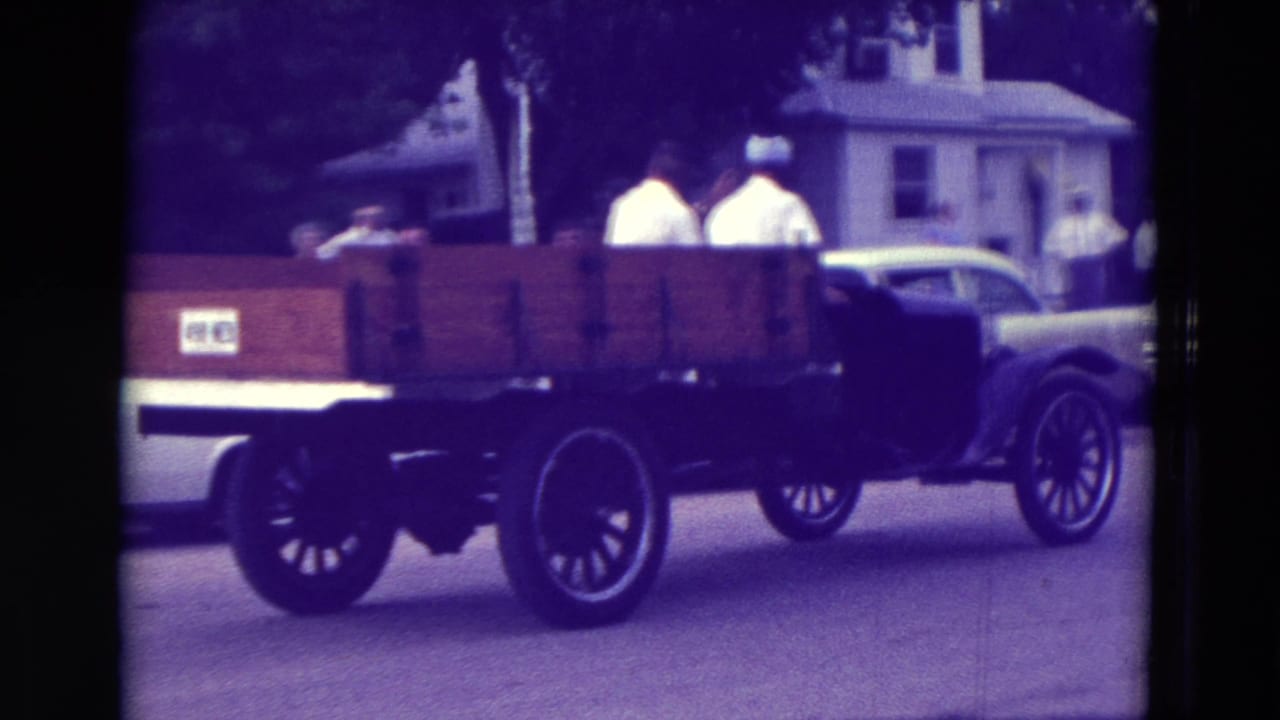 In Clarkson, Nebraska in 1965, an older wooden flatbed truck carries passengers past houses and parked cars during a parade.