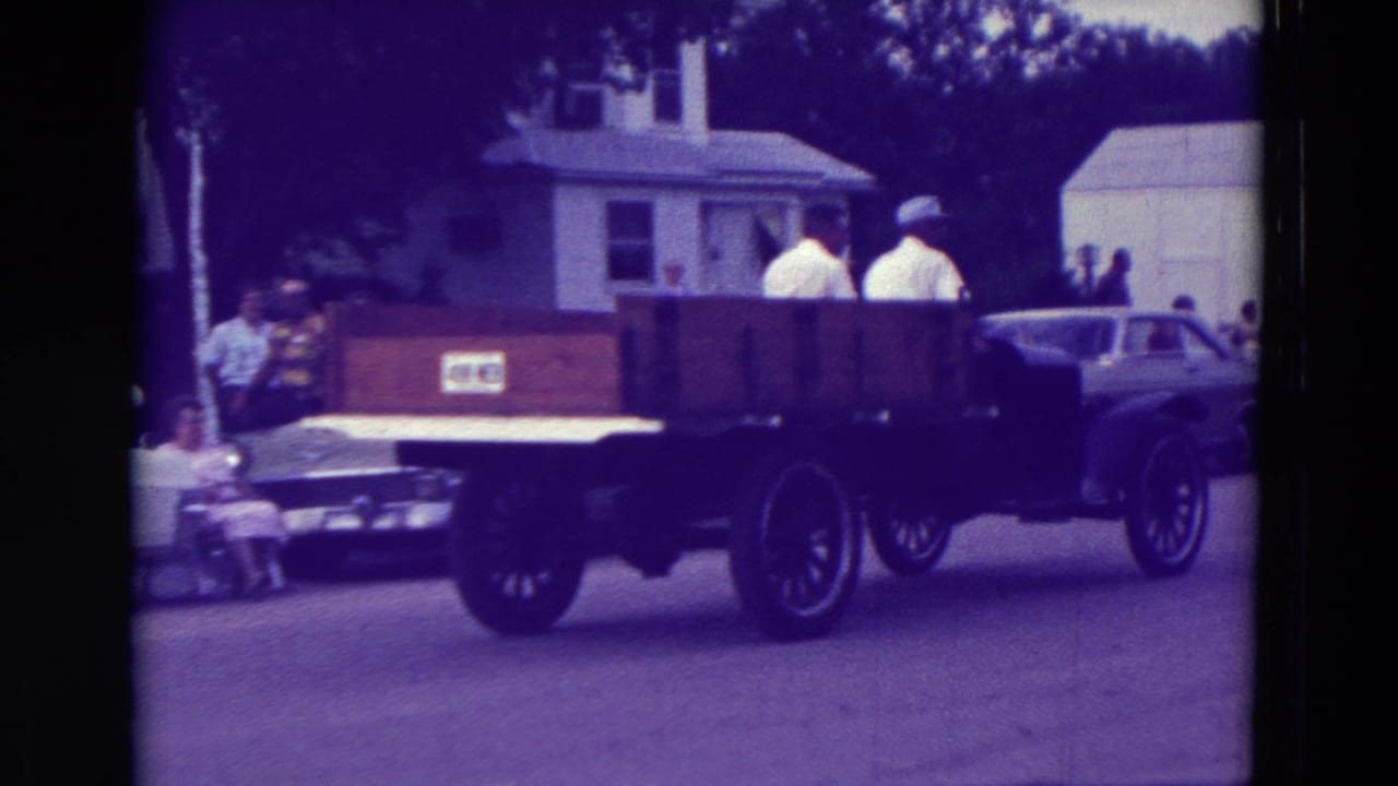 The wooden flatbed truck continues through the 1965 Clarkson, Nebraska parade with spectators and parked cars along the street.
