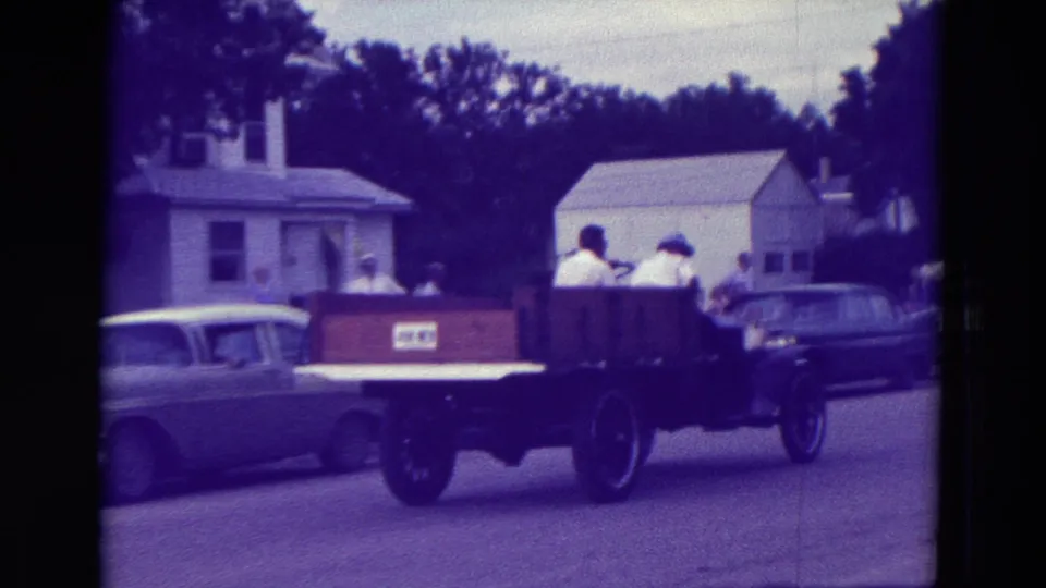 A rear view shows the wooden flatbed truck moving away during the 1965 Clarkson, Nebraska parade, with mid-century cars nearby.