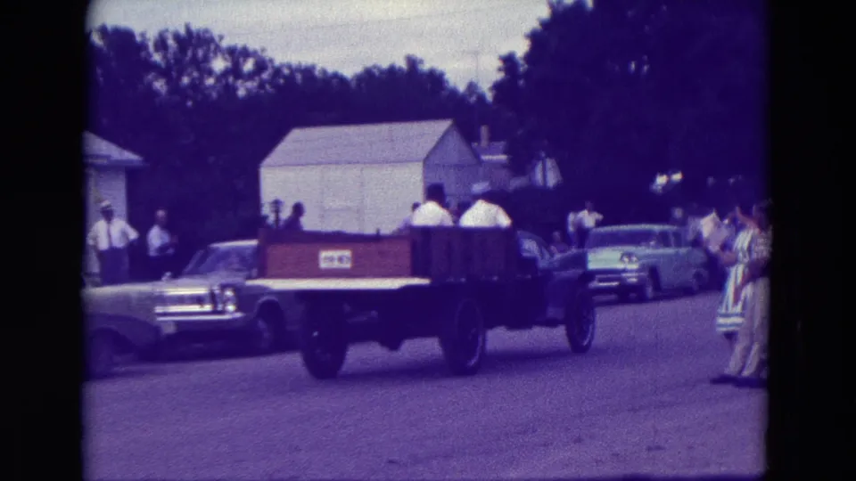 The 1965 Clarkson, Nebraska parade truck moves farther down the residential street as spectators and cars line the route.