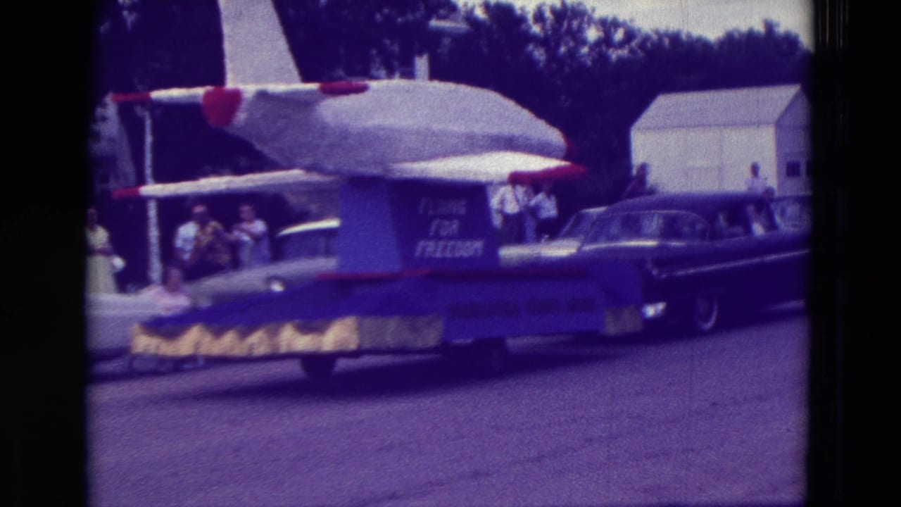 Airplane parade float pulled by a dark classic sedan with spectators along the street.
