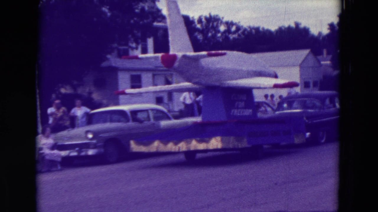 The airplane-themed parade float continues through Clarkson, Nebraska, with period cars, houses, and spectators visible behind it.