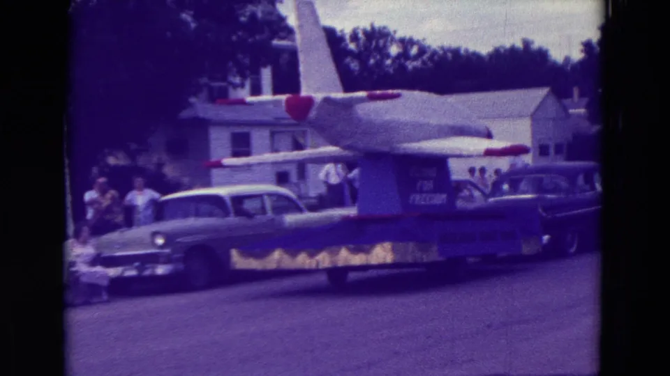 The airplane-themed parade float continues through Clarkson, Nebraska, with period cars, houses, and spectators visible behind it.