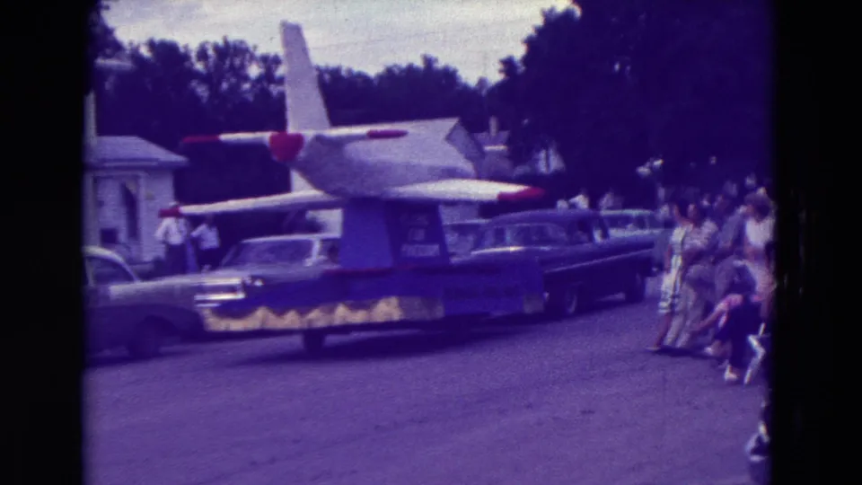The airplane float moves away along a Clarkson street while spectators in 1960s clothing watch from the curb.