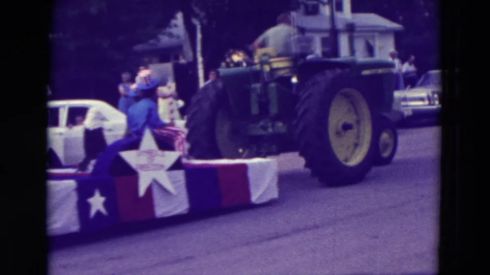 A green tractor pulls a red, white, and blue float with star decoration in the same Clarkson, Nebraska parade footage.