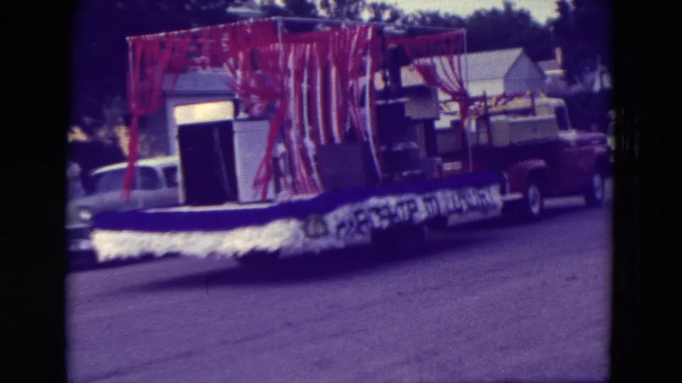 1965 Clarkson, Nebraska parade float receding past the camera, pulled by a red pickup with a white car behind it.