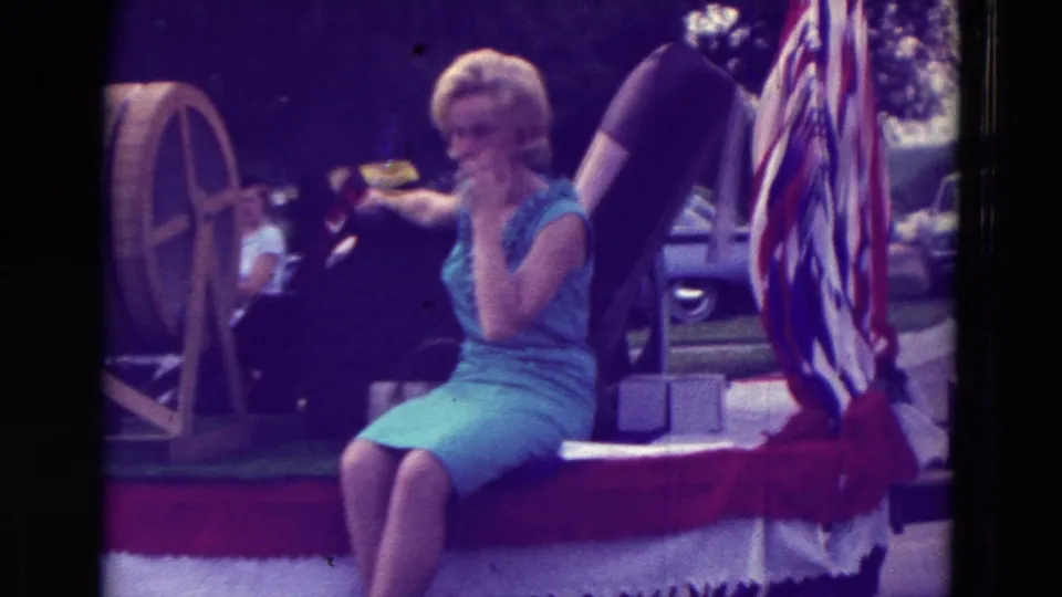 A blonde woman in a sleeveless blue dress rides on a patriotic parade float in Clarkson, Nebraska, in 1965.