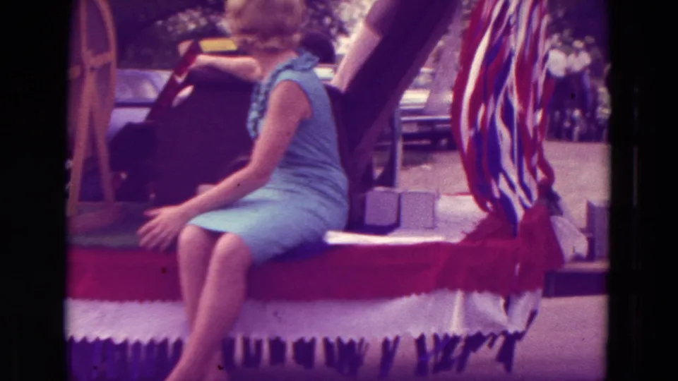 The home-movie frame shows the woman seated on the float edge with patriotic bunting and parade props around her.