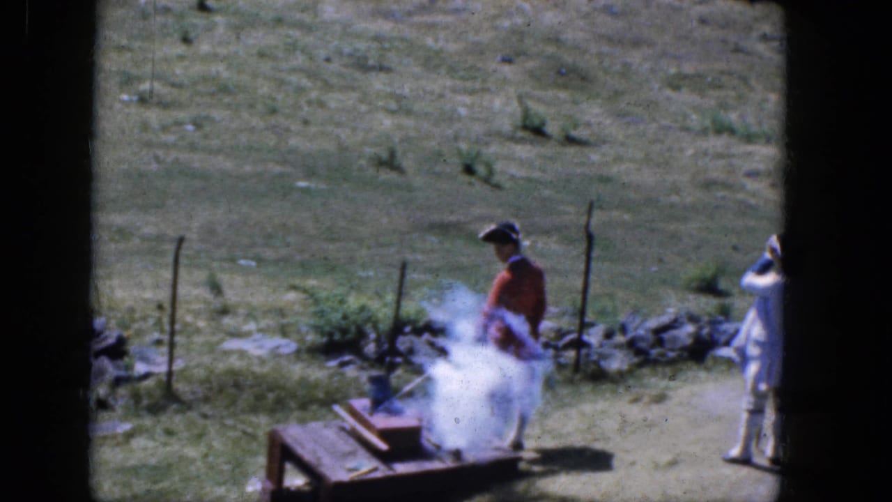 Two costumed reenactors stand near a smoky wooden platform in a grassy field.