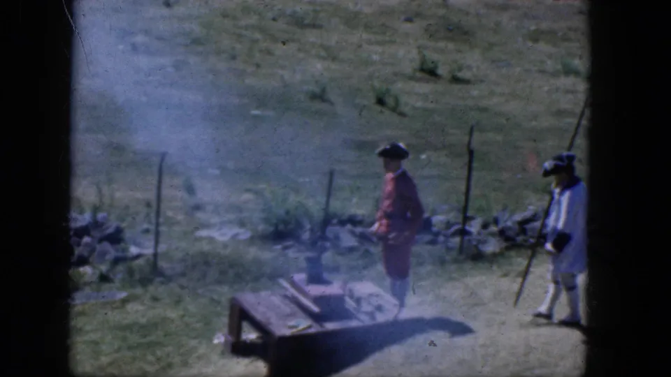 Two period-costumed reenactors appear near a smoky weapon demonstration in a grassy Concord field in 1965.