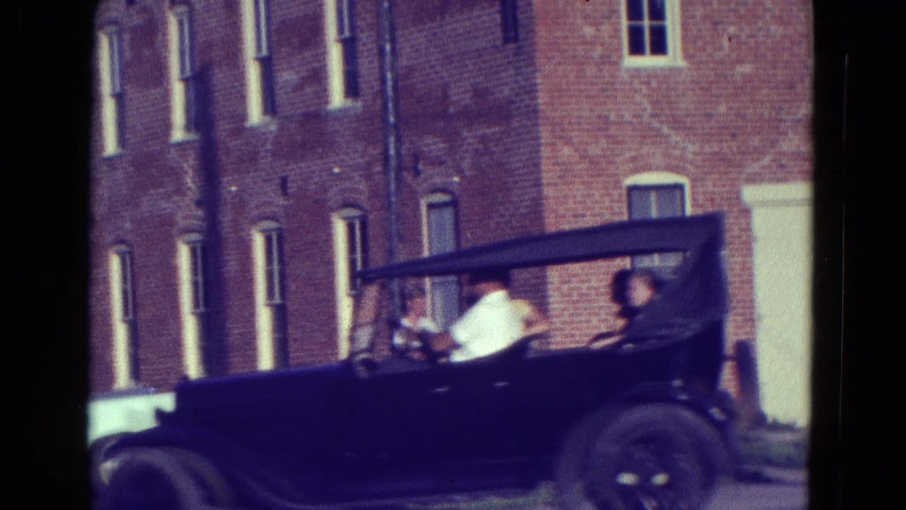 Dark soft-top vintage car passing a red brick building with passengers inside.
