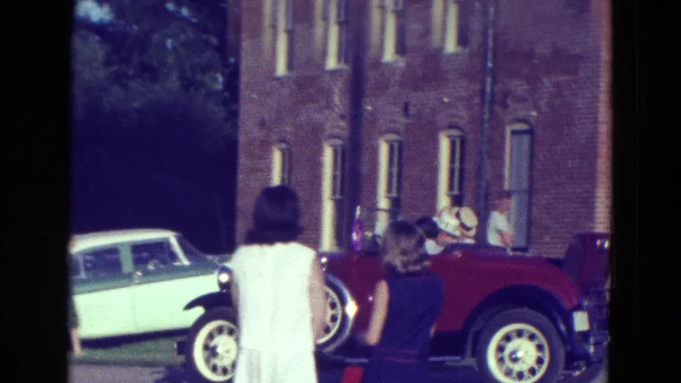 Red open-top car with passengers near a red brick building, with a foreground woman and a light-colored car in view.