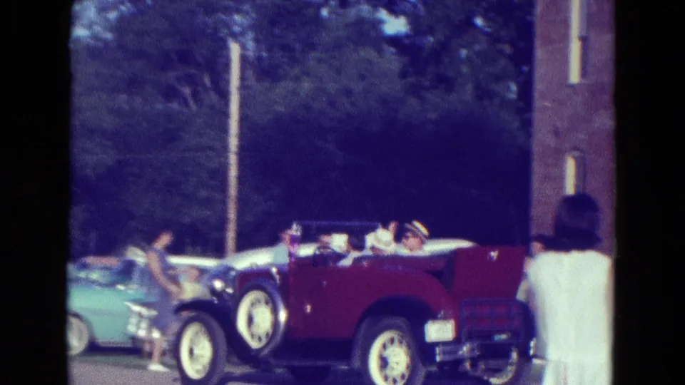 Red open-top car and light blue car appear beside a brick building in 1965 Diller, Nebraska, with several people visible around them.