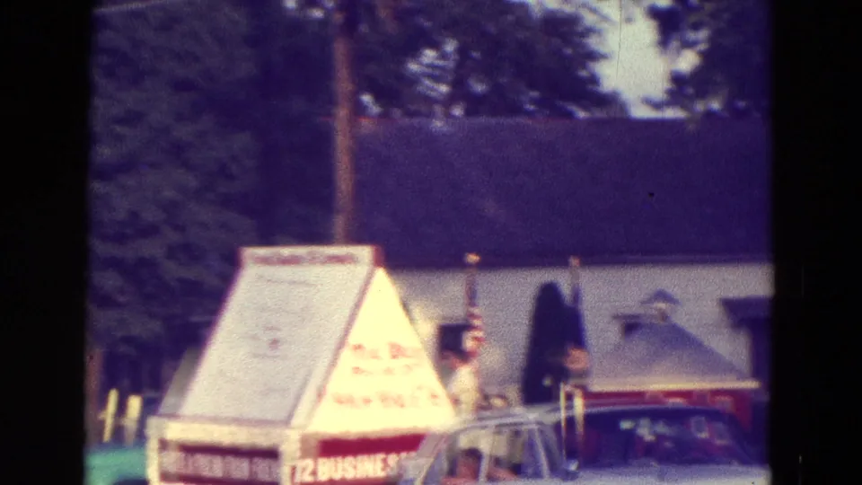 1965 Diller, Nebraska home-movie frame showing a triangular festival display with hand-painted text near classic cars and trees.