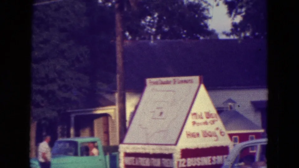 1965 small-town festival footage in Diller, Nebraska showing the Highway 6 display near a turquoise pickup, a car, and a spectator.