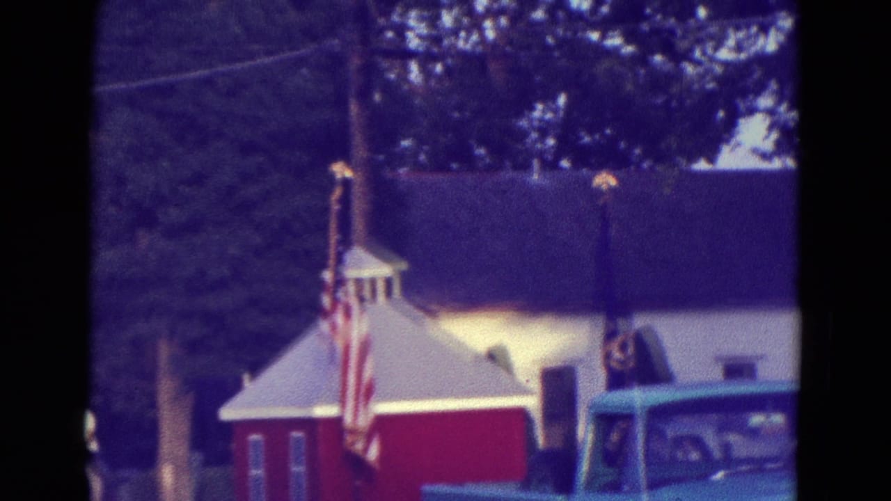 Red building with American flags and a partially visible turquoise vehicle on a tree-lined street.