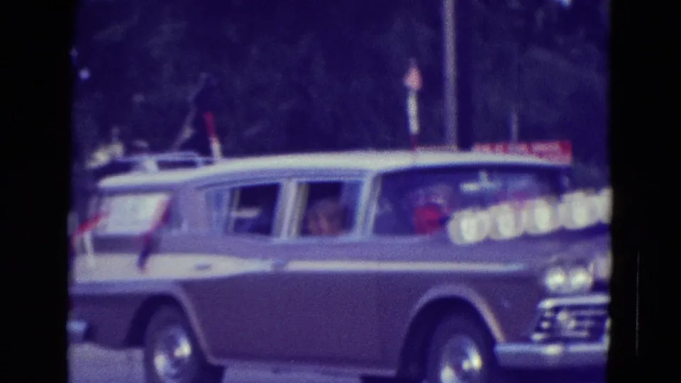 Light-colored station wagon decorated with small American flags moves through the 1965 Diller, Nebraska parade scene.