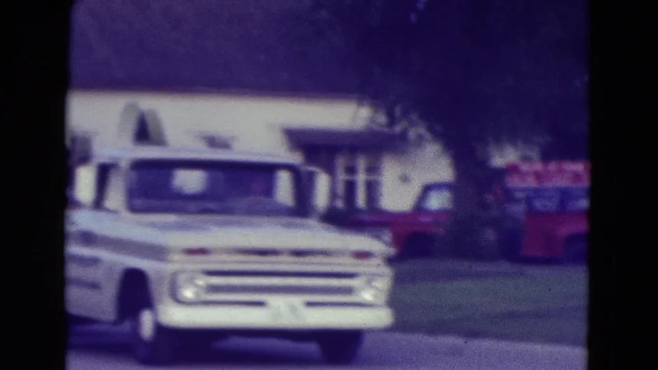White vintage pickup travels along the Diller, Nebraska street with a building, trees, and red vehicle behind it.
