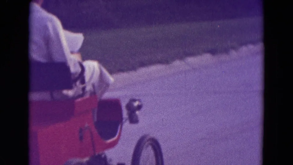A closer 1965 home-movie view shows the red go-kart front, wheel, and seated white-clad rider on the street.