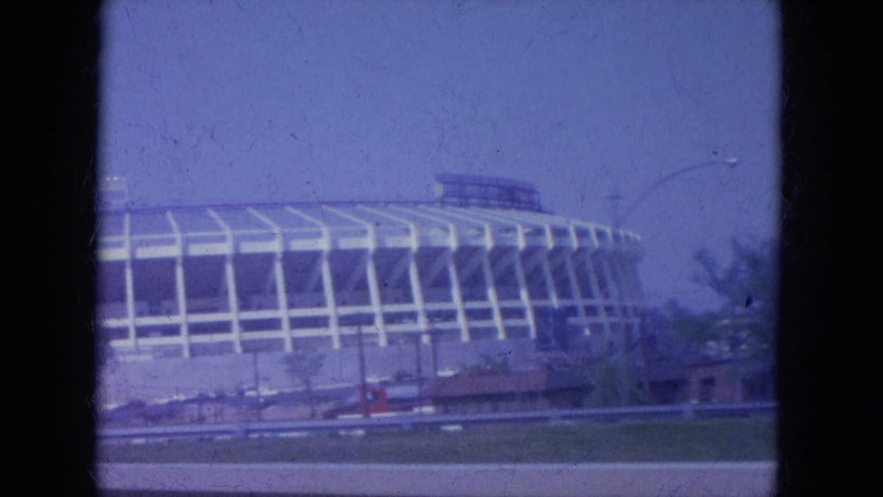 Large stadium exterior seen through an 8mm film frame from a moving vehicle.