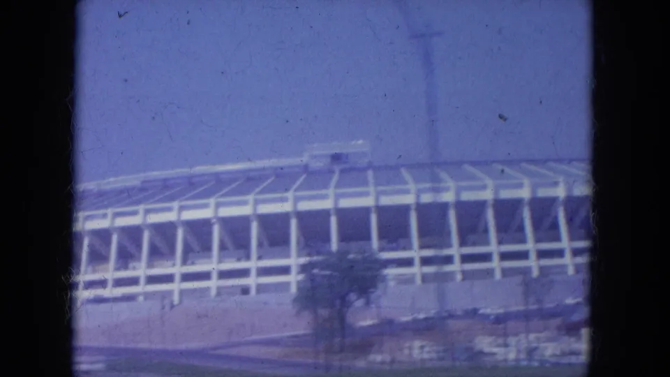 1965 Louisville stadium exterior viewed from a vehicle, showing white support ribs, a low wall, blue sky, and roadside landscaping.