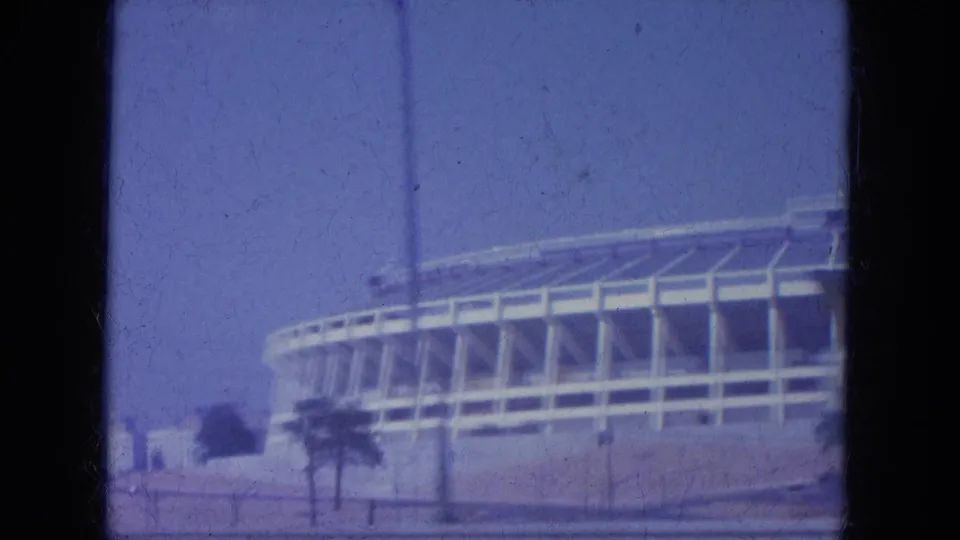 1965 Louisville drive-by frame showing the stadium's curved exterior, tall roadside poles, palm-like trees, and faded blue 8mm color.