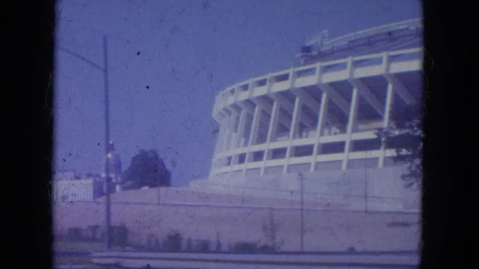 1965 Louisville stadium exterior at the end of a vehicle pass, with the structure partly cropped beside a wall, poles, trees, and faded sky.