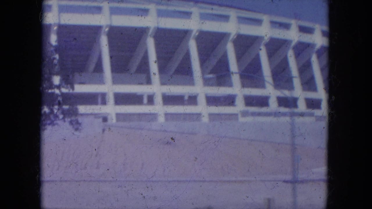 White stadium exterior with curved upper rim, support columns, concrete wall, and blue sky.