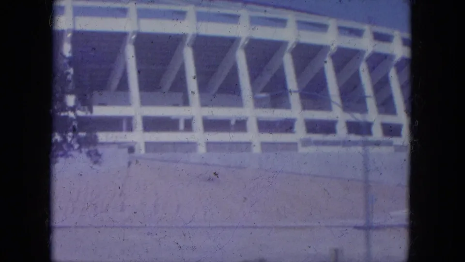1965 Louisville home-movie frame showing a white stadium exterior with angled supports, concrete base, and blue sky.