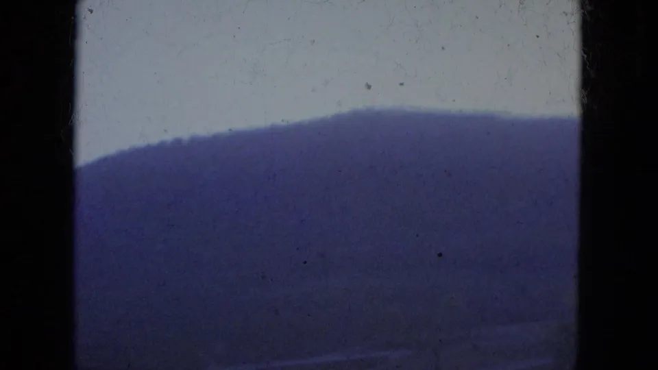 Soft 8mm travel view of a dark wooded hillside and pale sky, with no people or signs visible.