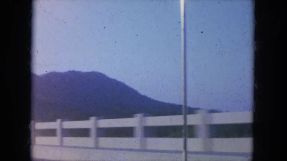 Passenger-view 8mm frame of a bridge railing, vertical pole, and hills during a 1965 Louisville-area drive.