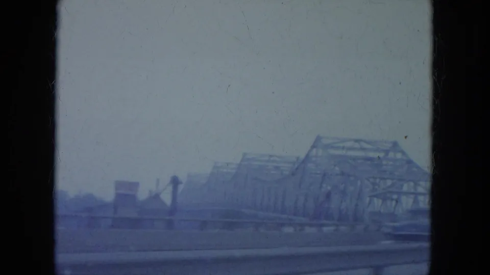 1965 Louisville home-movie frame showing a roadway view toward steel truss bridge spans under an overcast sky.