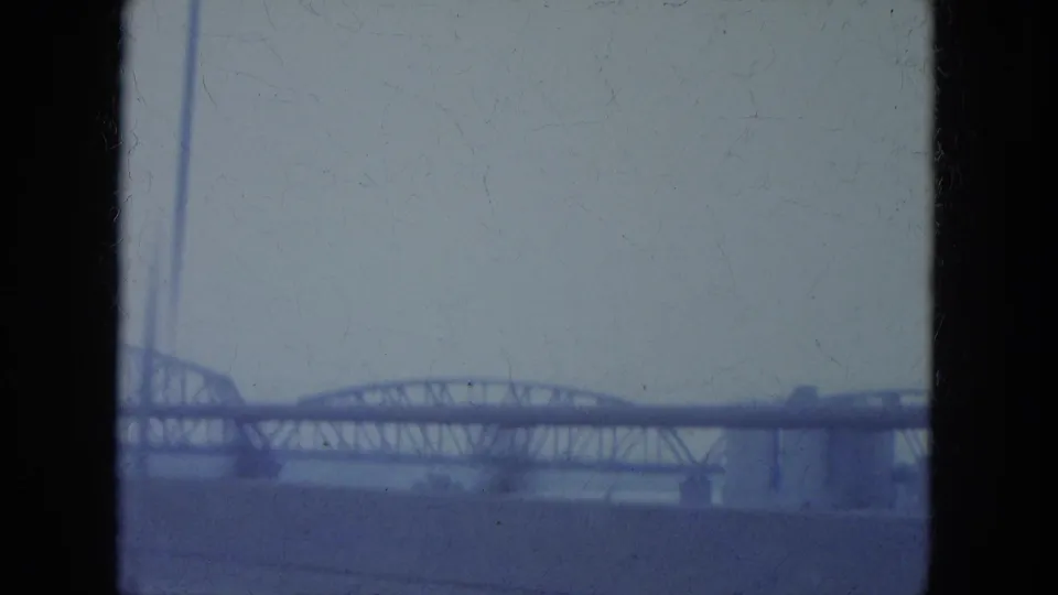 1965 Louisville home-movie frame showing a distant steel truss bridge and roadway approach with dark film borders.