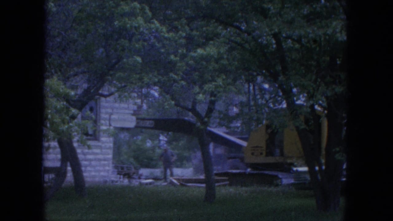 Yellow backhoe beside a stone building behind trees.