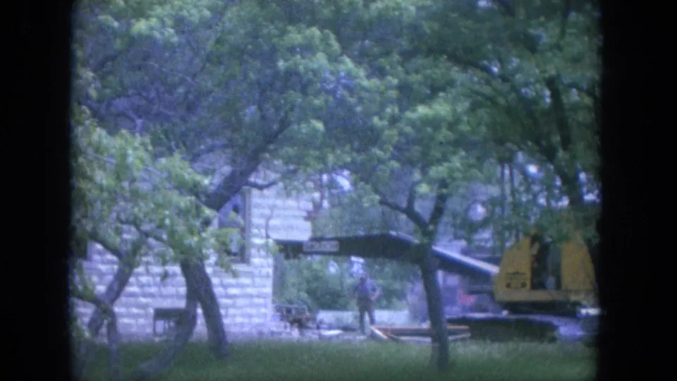 1965 Louisville demolition view with a yellow backhoe positioned by a stone building under dense tree cover.