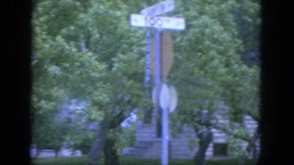 1965 Louisville frame showing a street sign pole and dense trees with the stone building partly visible behind them.