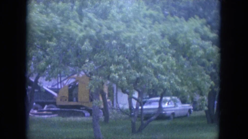1965 Louisville street scene with a yellow construction vehicle, parked car, trees, grass, and a house in the background.